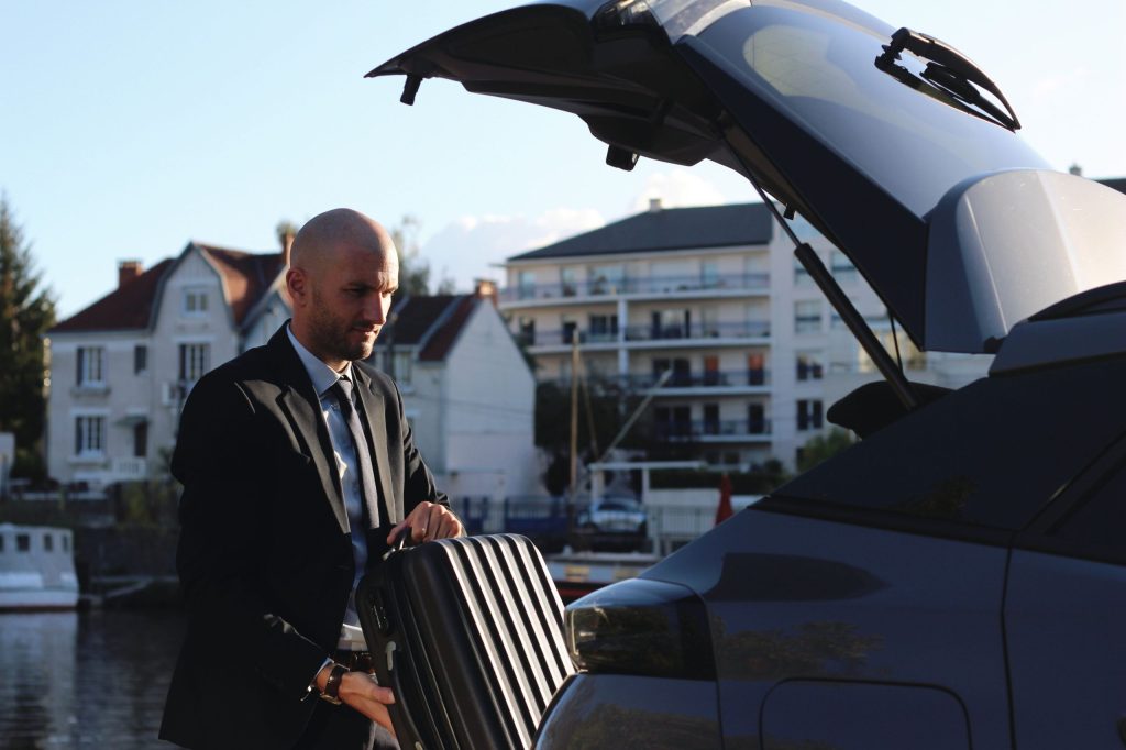 Un chauffeur en costume noir et cravate plaçant une valise à rayures noires dans le coffre ouvert d'un SUV, avec en arrière-plan des maisons blanches et un bord de l'eau, probablement l'Erdre à Nantes.