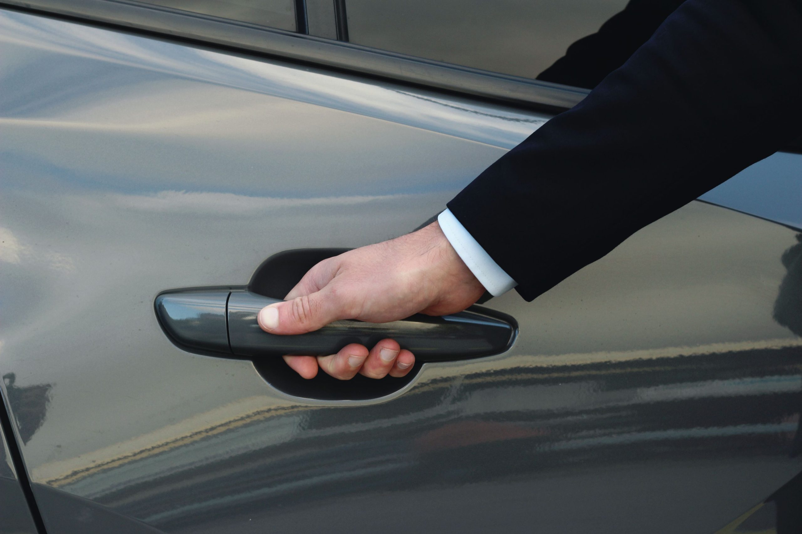 Gros plan sur la main d'un chauffeur en costume noir et chemise blanche ouvrant la poignée d'une portière de voiture grise.