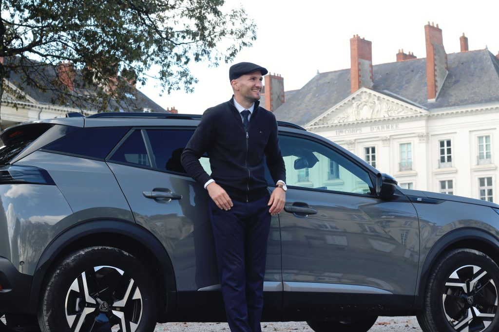 Un chauffeur souriant en costume et casquette appuyé contre son SUV Peugeot gris, devant un bâtiment historique nantais avec fronton sculpté et inscription "Corps d'Armée".