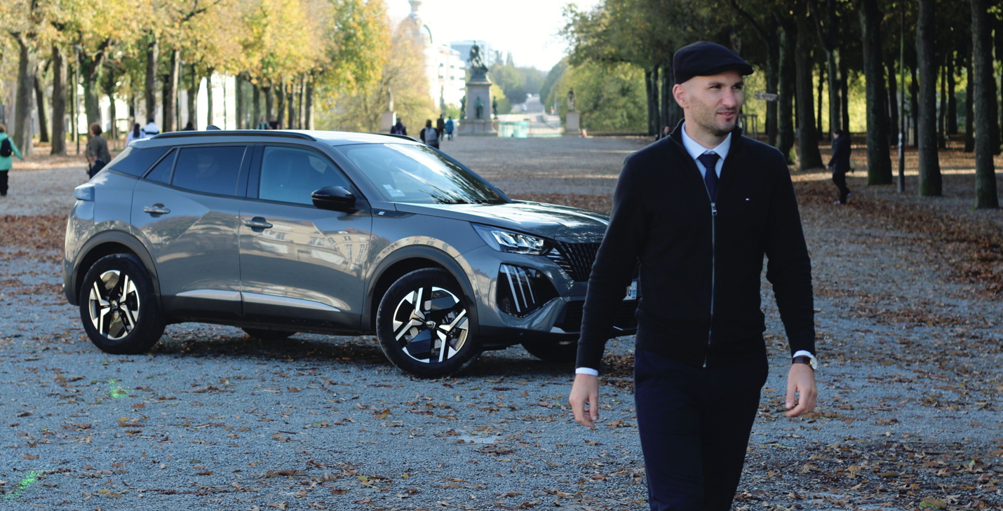 Un chauffeur en costume et casquette marchant devant son SUV Peugeot gris garé dans le Cours Saint-Pierre à Nantes en automne, avec des allées d'arbres aux feuilles dorées et une statue visible en arrière-plan.