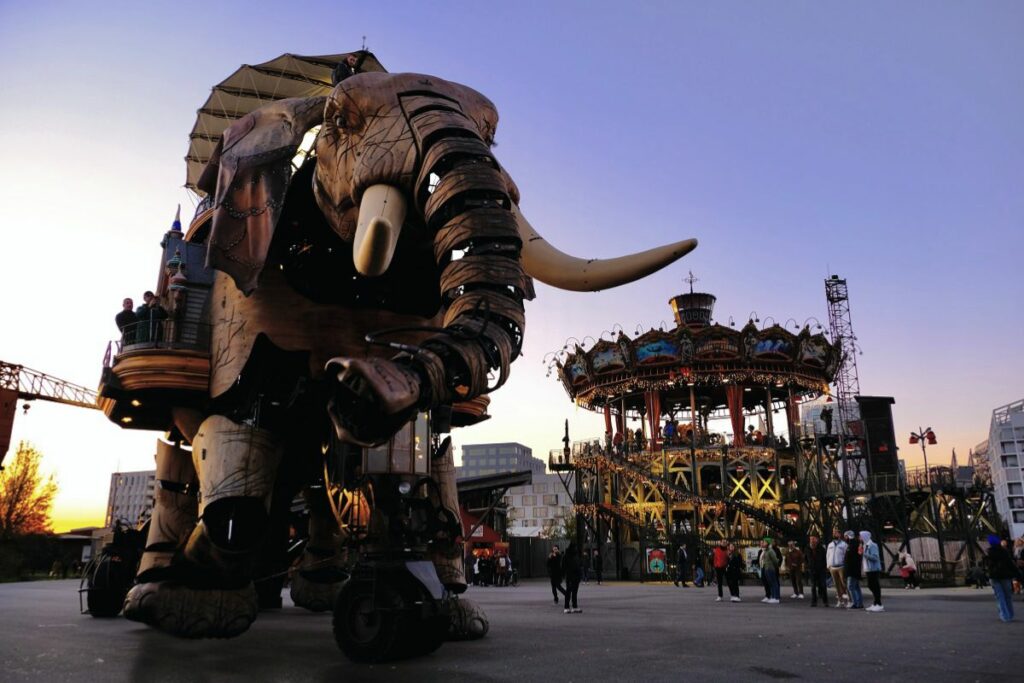 Le Grand Éléphant mécanique des Machines de l'île de Nantes au coucher du soleil, avec le carrousel des Mondes Marins (inspiré par l'univers de Jules Verne) à plusieurs étages visible en arrière-plan et des visiteurs qui admirent ces structures monumentales.