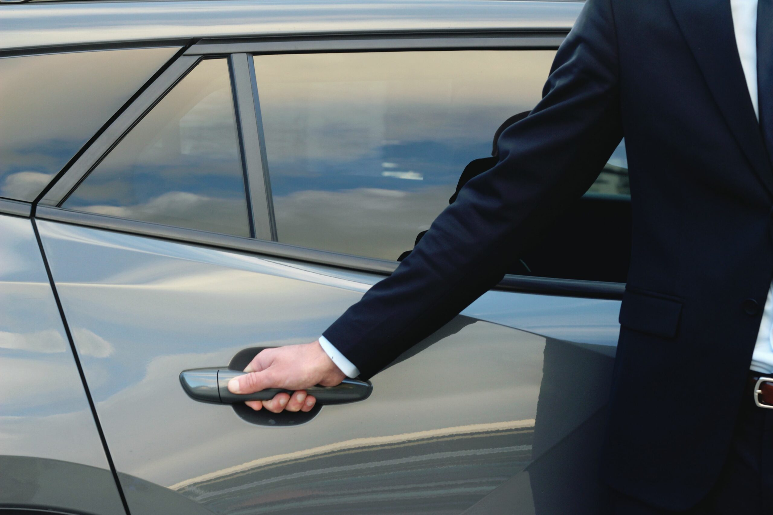 Un chauffeur en costume élégant ouvrant la porte arrière d'une berline haut de gamme, montrant le bras en costume sombre et manchette blanche saisissant la poignée de porte.
