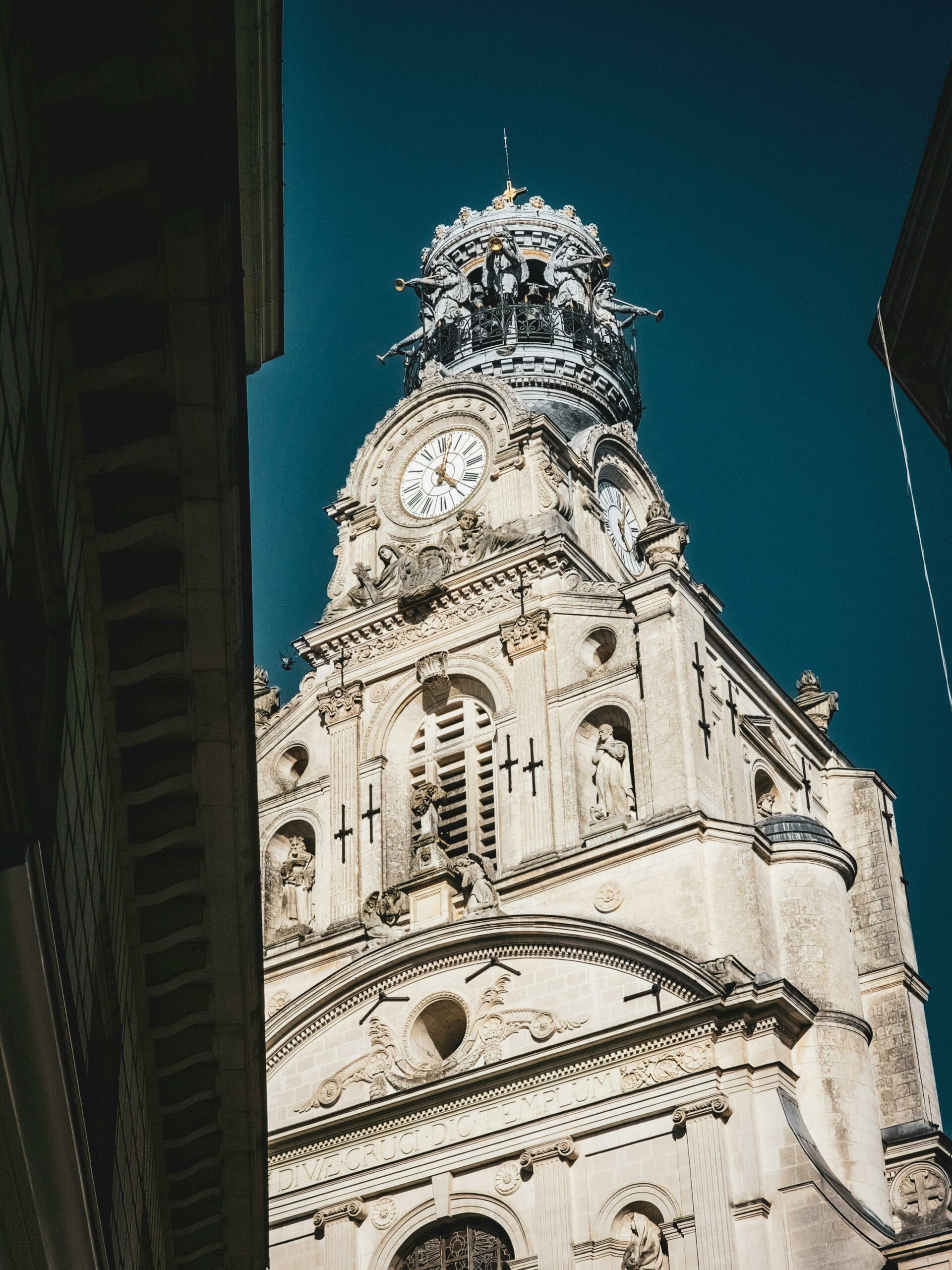 Vue en contre-plongée du beffroi de l'église Sainte-Croix à Nantes, montrant sa tour clocher en pierre blanche ornée de sculptures, avec son horloge et son dôme décoratif, se détachant sur un ciel bleu profond.