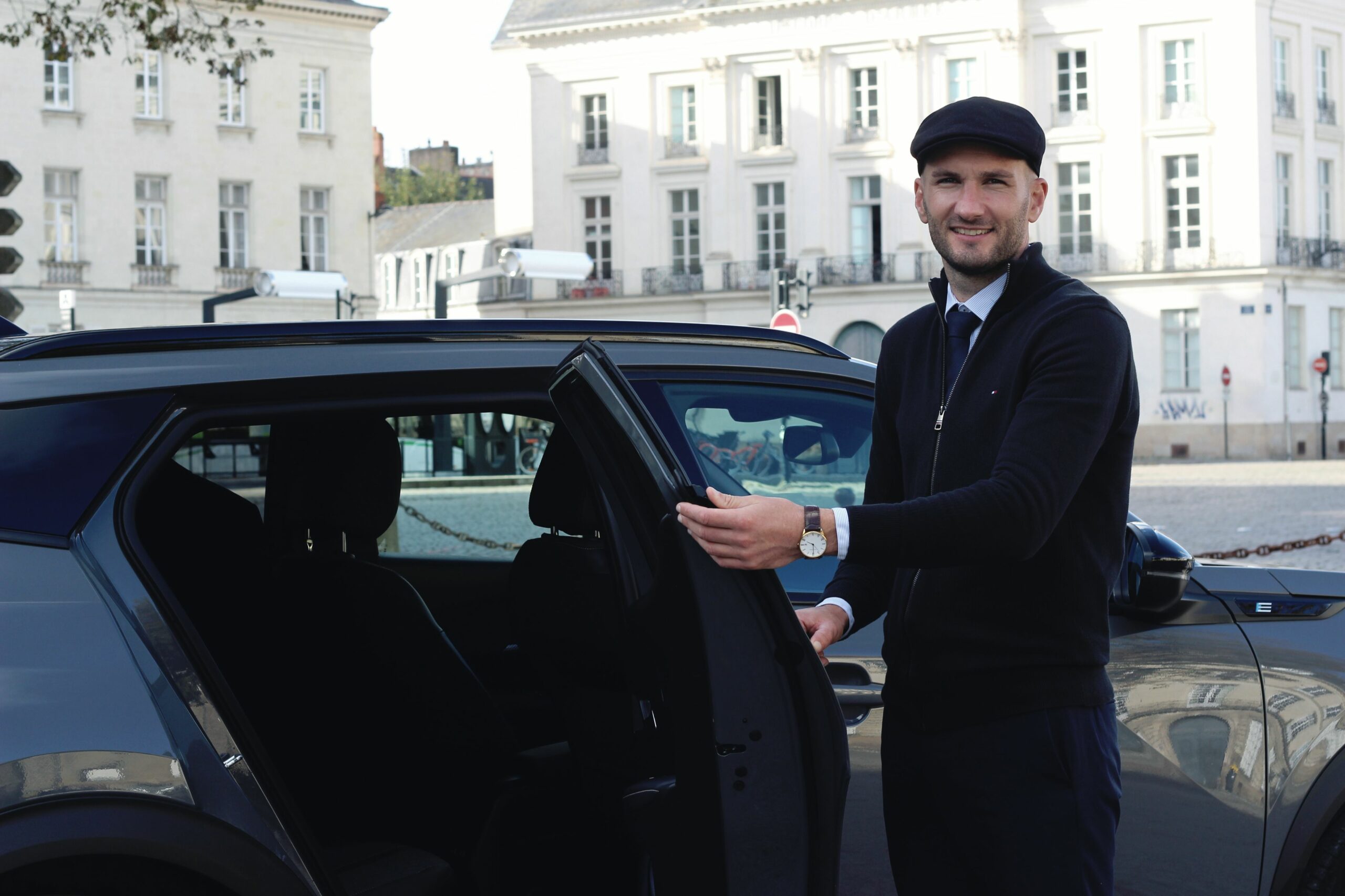 Un chauffeur élégant et souriant ouvre la portière d'une voiture de luxe devant l'Hôtel d'Aux, ancien siège du XIe corps d'armée longeant le cours Saint-André à Nantes.
